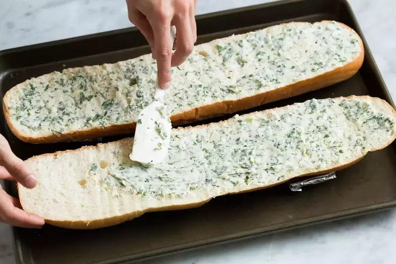 A person spreading garlic butter onto bread slices before toasting them in the oven.
