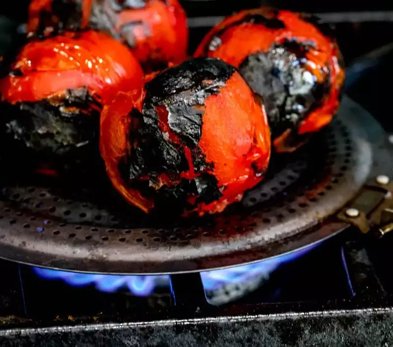Close-up of juicy tomatoes roasting on an open flame, with charred skin beginning to form.